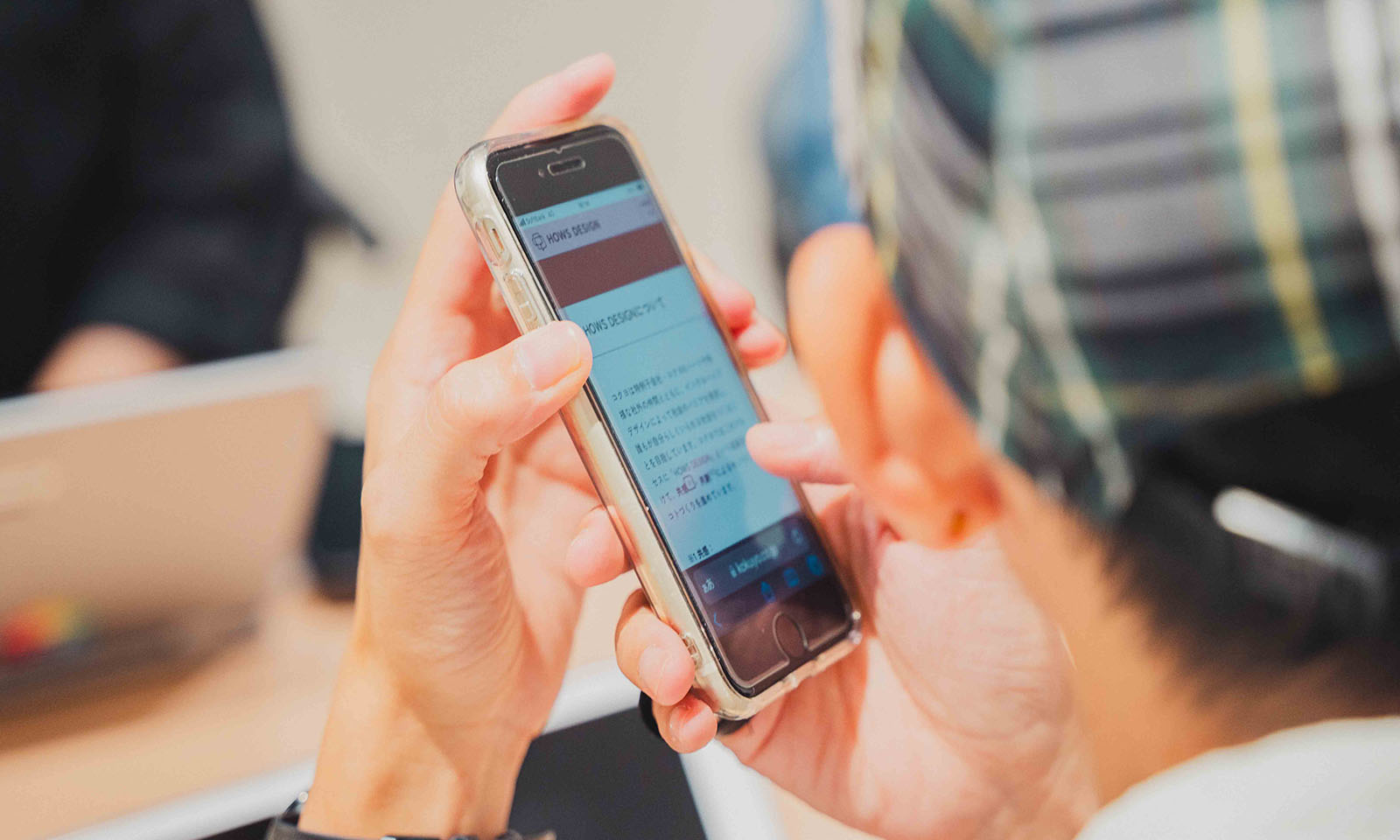 ALTはじまり。チェックの帽子を被った石井さんがiPhoneのスクリーンリーダーでHOWS DESIGNのWEBサイトを開き、少し耳のそばに近づけて内容を聞いている様子。ALTおわり