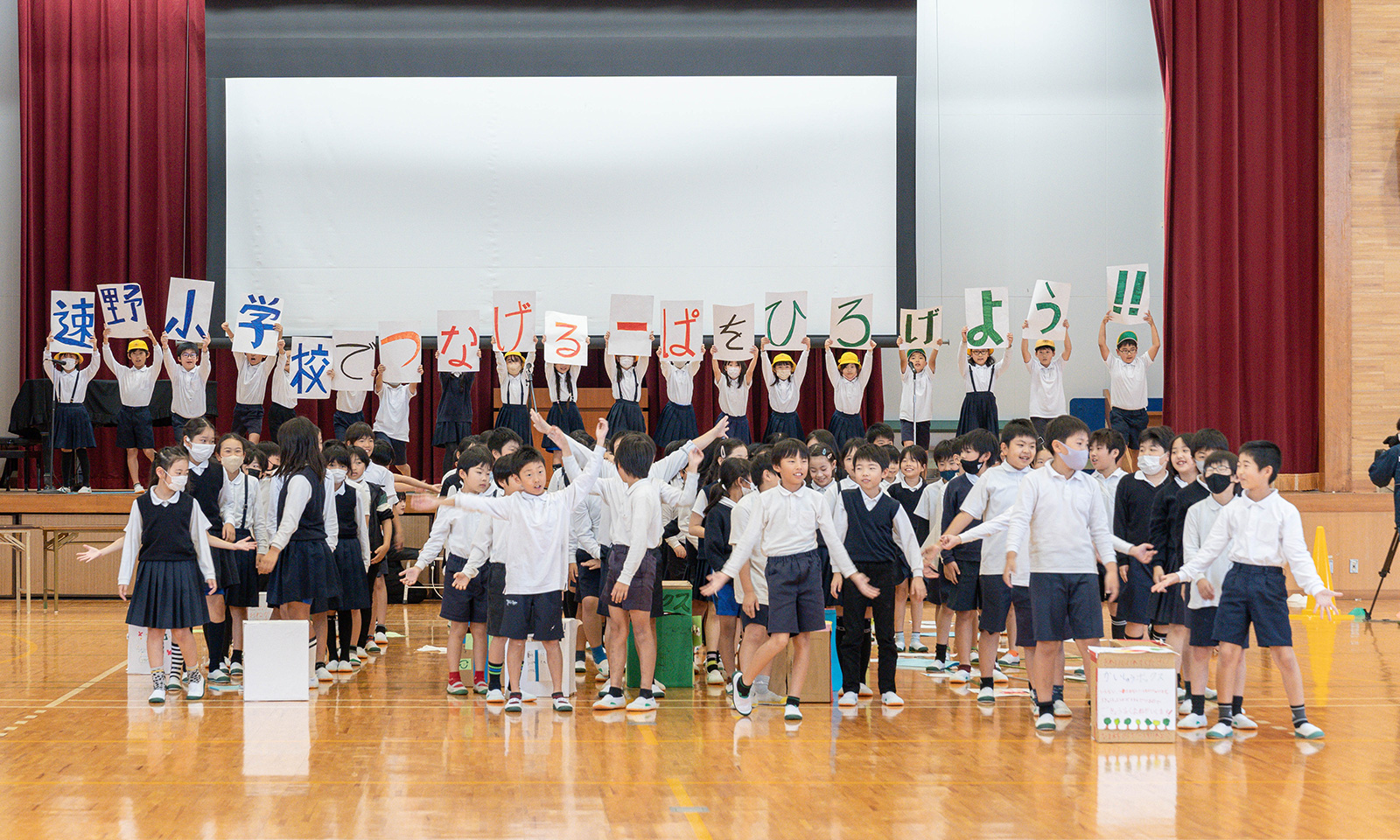 全校集会で、4年生たちが体育館の舞台の上で、「速野小学校でつなげるーぱをひろげよう!!」というプラカードを高く掲げている。舞台に上がっていない4年生たち、約50名も大きく横に手を広げ、全校生徒に向けて呼びかけている様子。